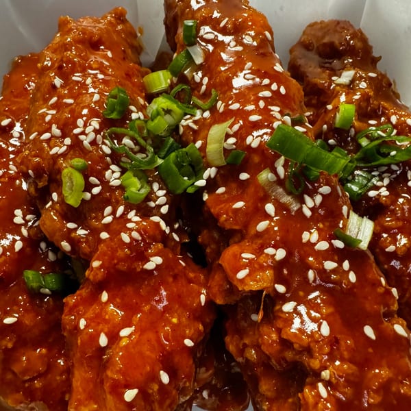Close-up of So-Ju Chikn' Korean fried chicken coated in red sauce, sesame seeds, and green onions.
